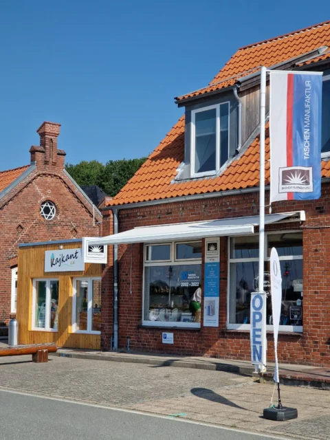 Außenansicht der Taschenmanufaktur Bootsmann am Tiessenkai in Kiel-Holtenau: Ein Ladengeschäft mit großen Schaufenstern und handgefertigten Taschen aus Segeltuch und LKW-Plane, gelegen direkt am Wasser des Tiessenkai mit Blick auf die Kieler Innenförde