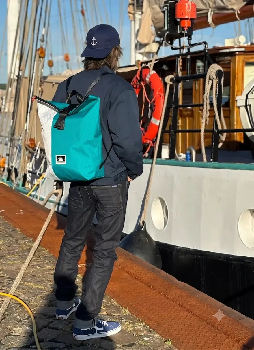 Firmengründer von Bootsmann Taschen steht am Tiessenkai in Kiel mit einem handgefertigten X-Pac-Rucksack an der Kaikante. Der wasserdichte Rolltop-Rucksack aus robustem Segellaminat wird vor maritimer Hafenkulisse präsentiert.