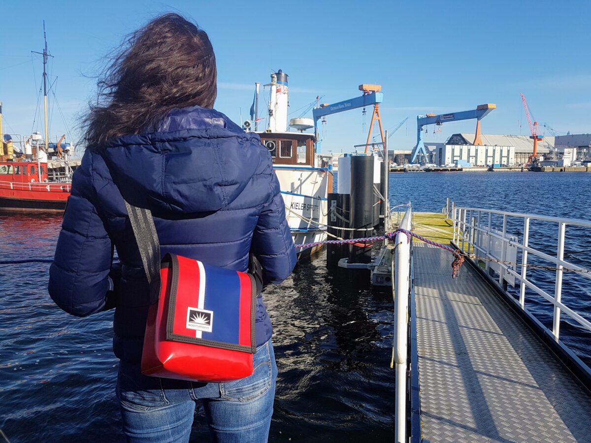 S-Tasche aus robuster LKW-Plane in Blau-Rot, getragen von Frau am Hafen vor urbaner Skyline