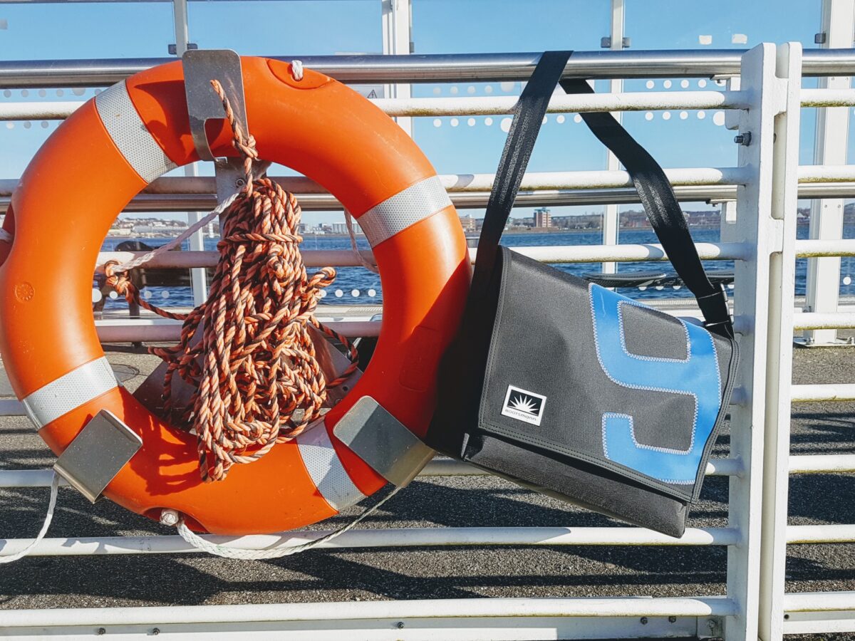 Große XL-Tasche aus Segeltuch hängt lässig an einem Hafen-Geländer neben einem orangen Rettungsring