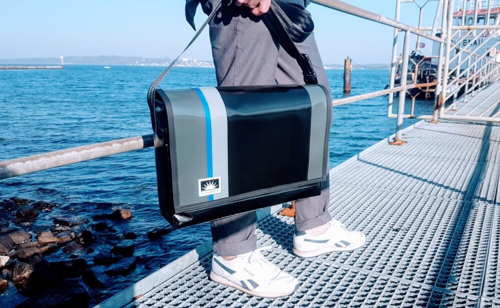 Laptoptasche aus LKW-Plane von Bootsman in 17 Zoll Größe, Mann mit Tasche an Geländer einer Seebrücke