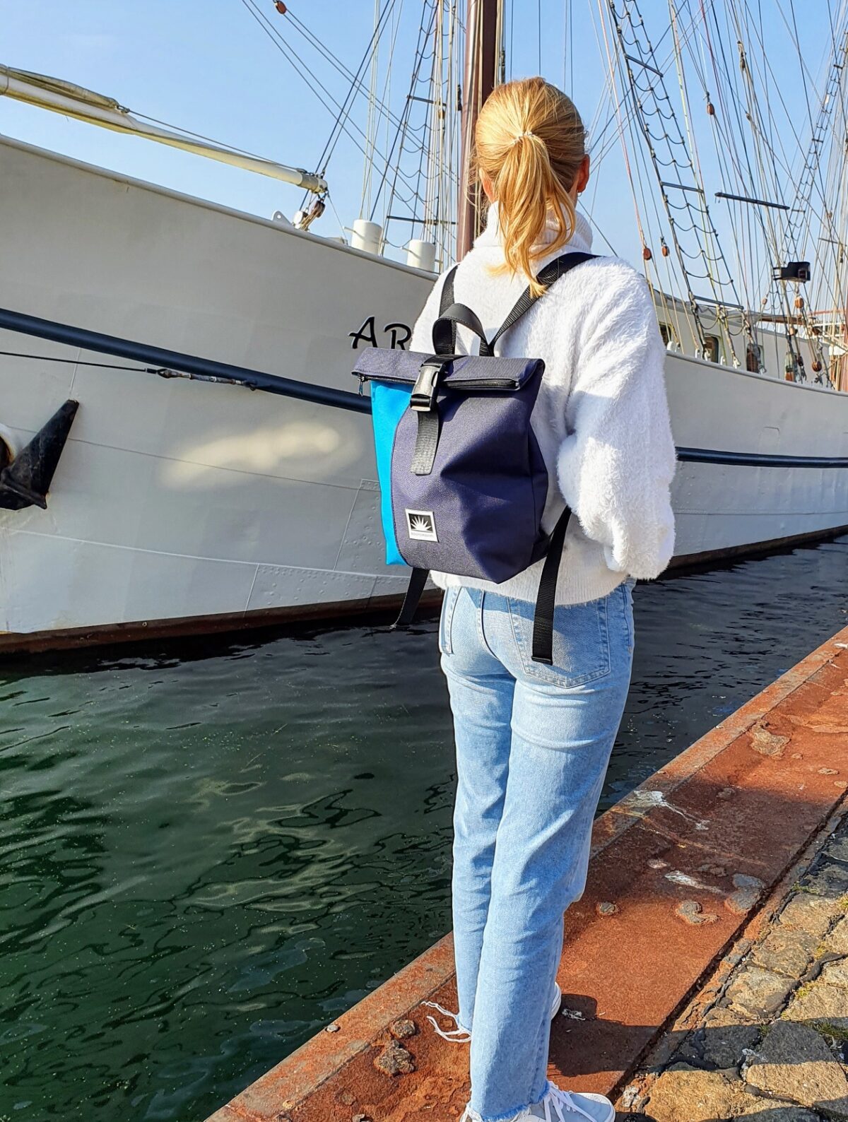 Frau steht am Kai, trägt einen BOOTSMANN-Rucksack aus Canvas in Himmelblau und Navyblau, Größe S, und schaut einem vorbeifahrenden Segelschiff hinterher