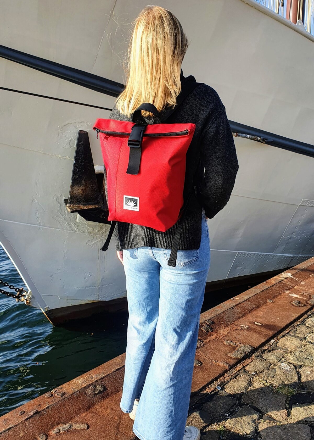 Junge Frau steht mit einem BOOTSMANN-Rucksack aus Canvas in Rot und Dunkelrot, Größe S, an der Kaikante, während ein altes Segelschiff ablegt