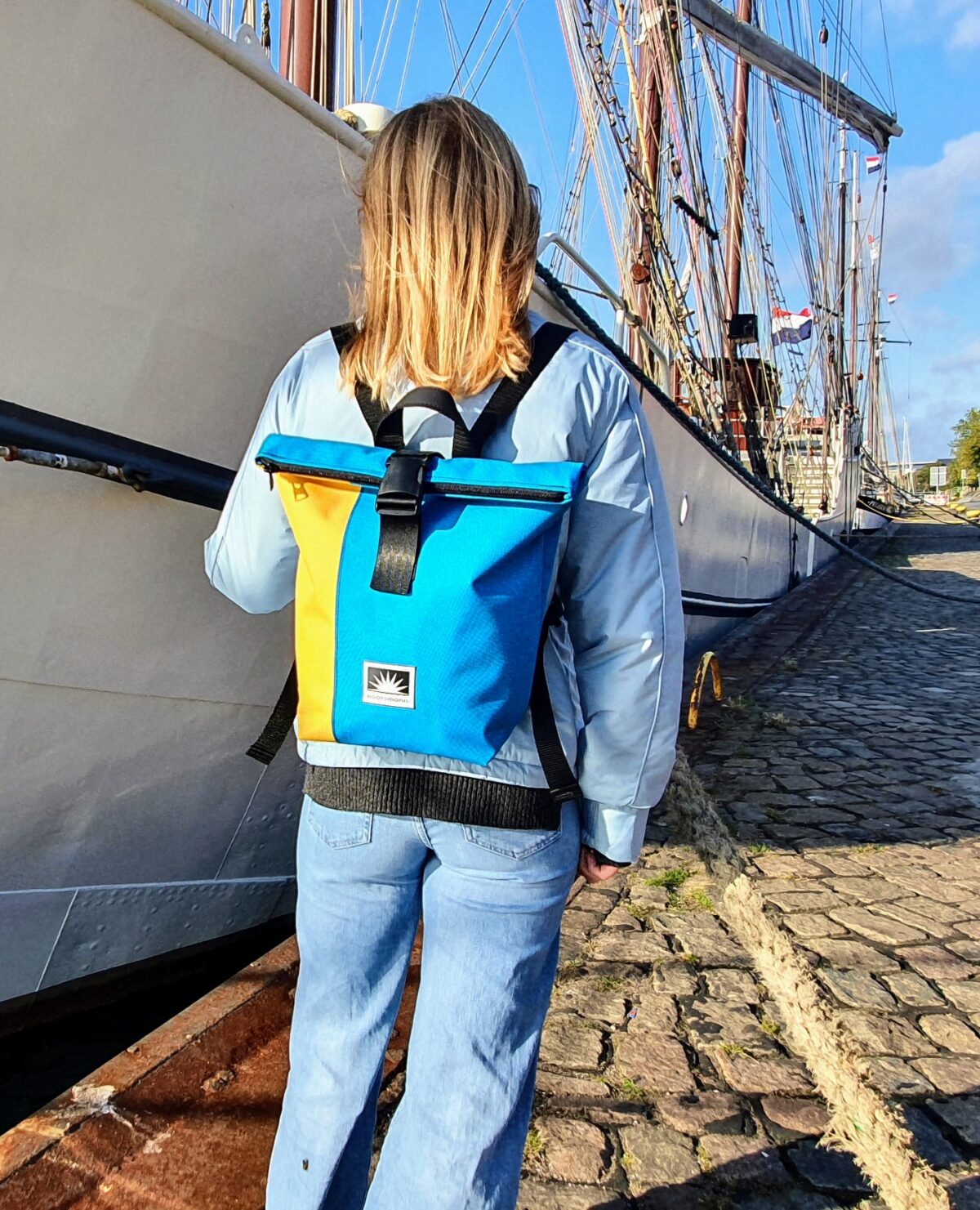 Frau steht am Kai, trägt einen BOOTSMANN-Rucksack aus Canvas in Gelb und Himmelblau, Größe S, und blickt auf mehrere Dreimaster im Hafen